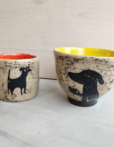 Photograph of two mugs. The first one is orange on the inside with a dog drawn on the outside; the second one is yellow on the inside with a dog holding a mug drawn on the outside.